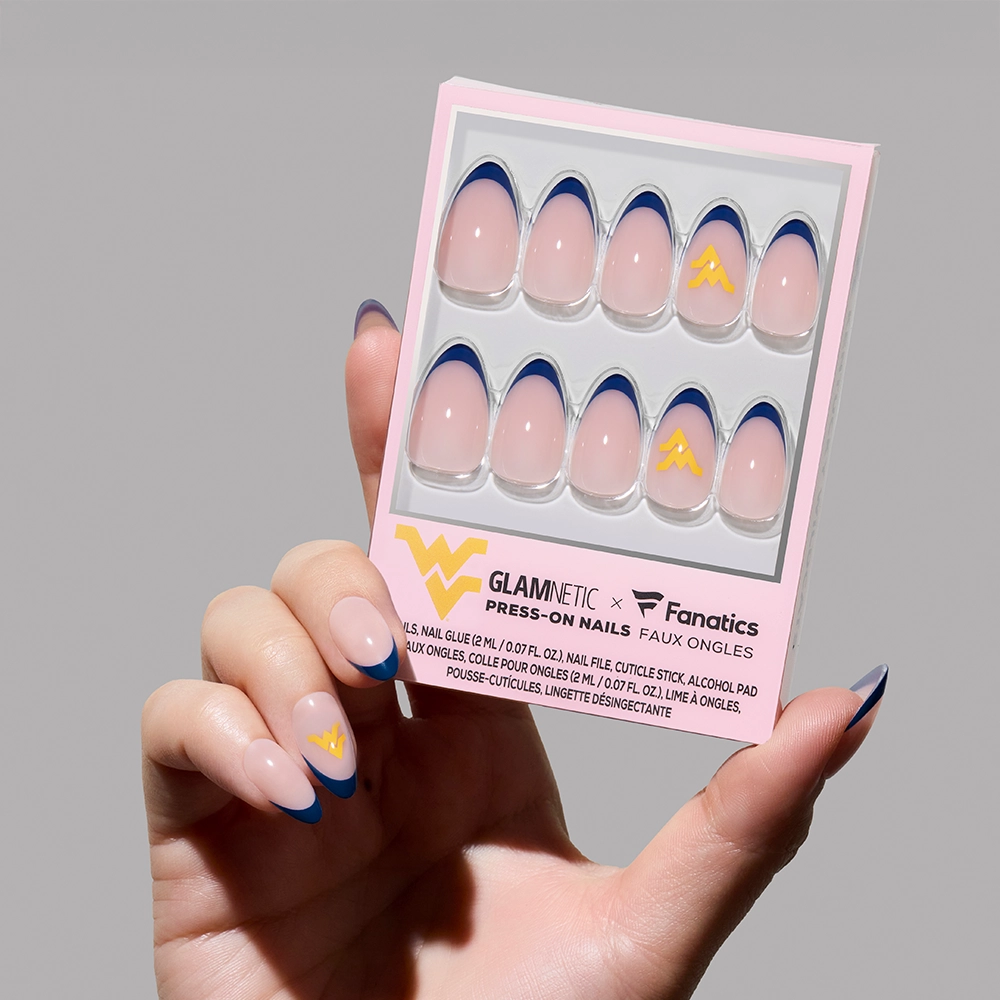 A hand with long, almond-shaped nails holds the glamnetic West Virginia Mountaineers Press-On Nail Set, featuring blue French tips and a yellow W logo—perfect for game day. The box is labeled "glamnetic x Fanatics. - Product image