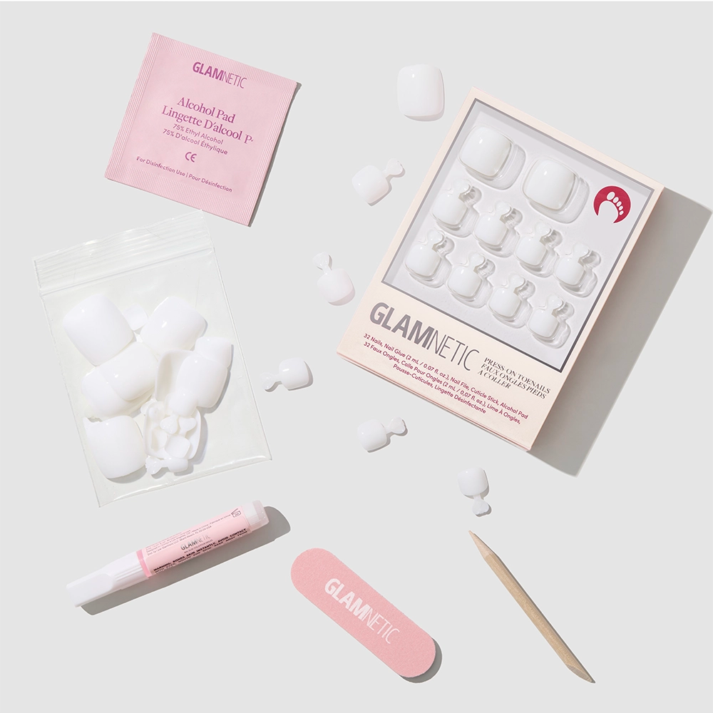 A flat lay of the glamnetic White Swan Toenail kit shows glossy press-on toenails, a pink nail file, glue tube, alcohol pad, wooden cuticle stick, and extra nails arranged on a light background. - Product image