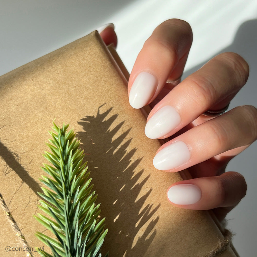 A hand with neatly manicured, short oval nails in glamnetics White Swan shade holds a brown paper-wrapped gift decorated with a pine branch; sunlight highlights the nails’ glossy finish. - Product image