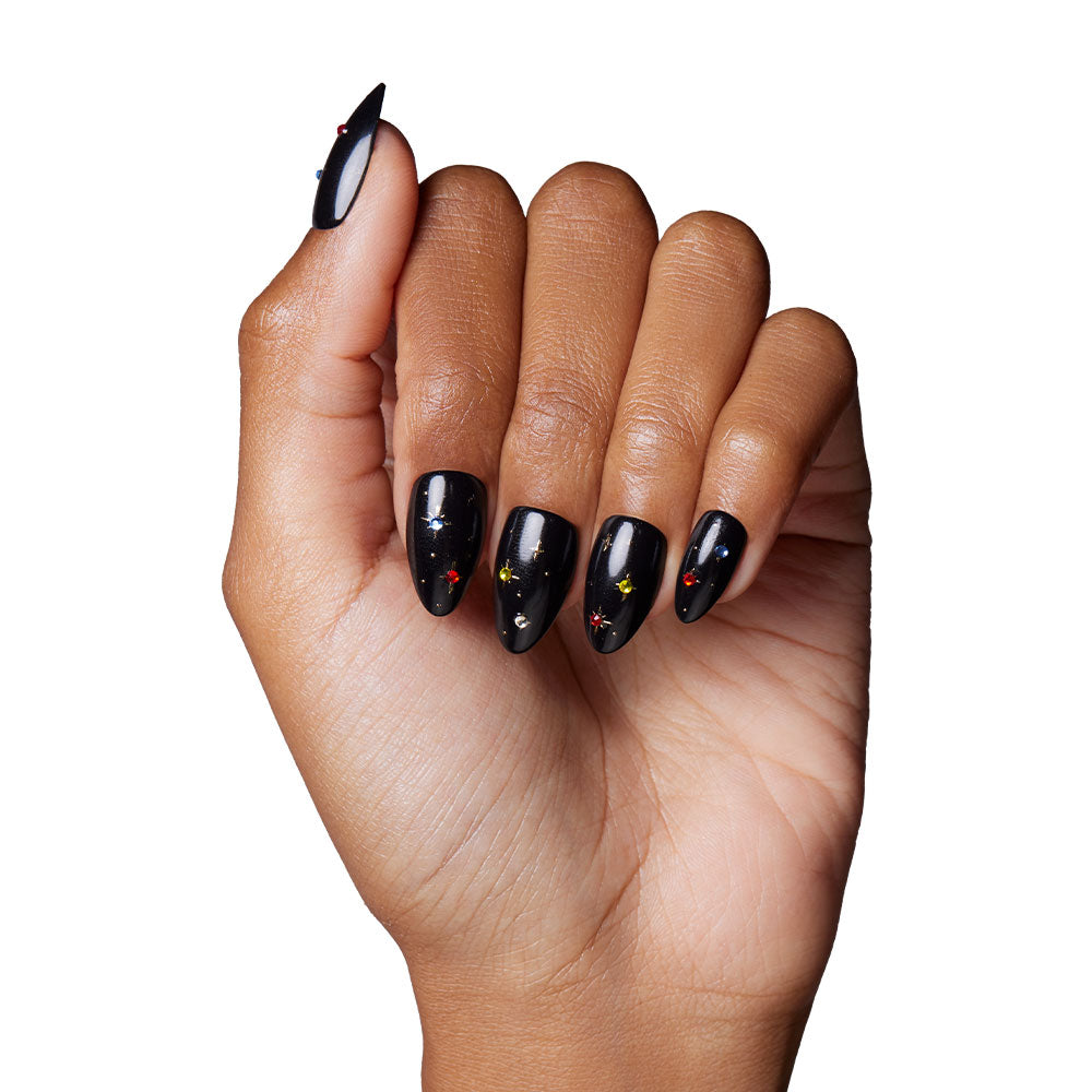 A hand with medium brown skin displays long, almond nails in glamnetics Winter Comet—glossy black with red and yellow gem accents—evoking celestial glam against a white background. - Product image