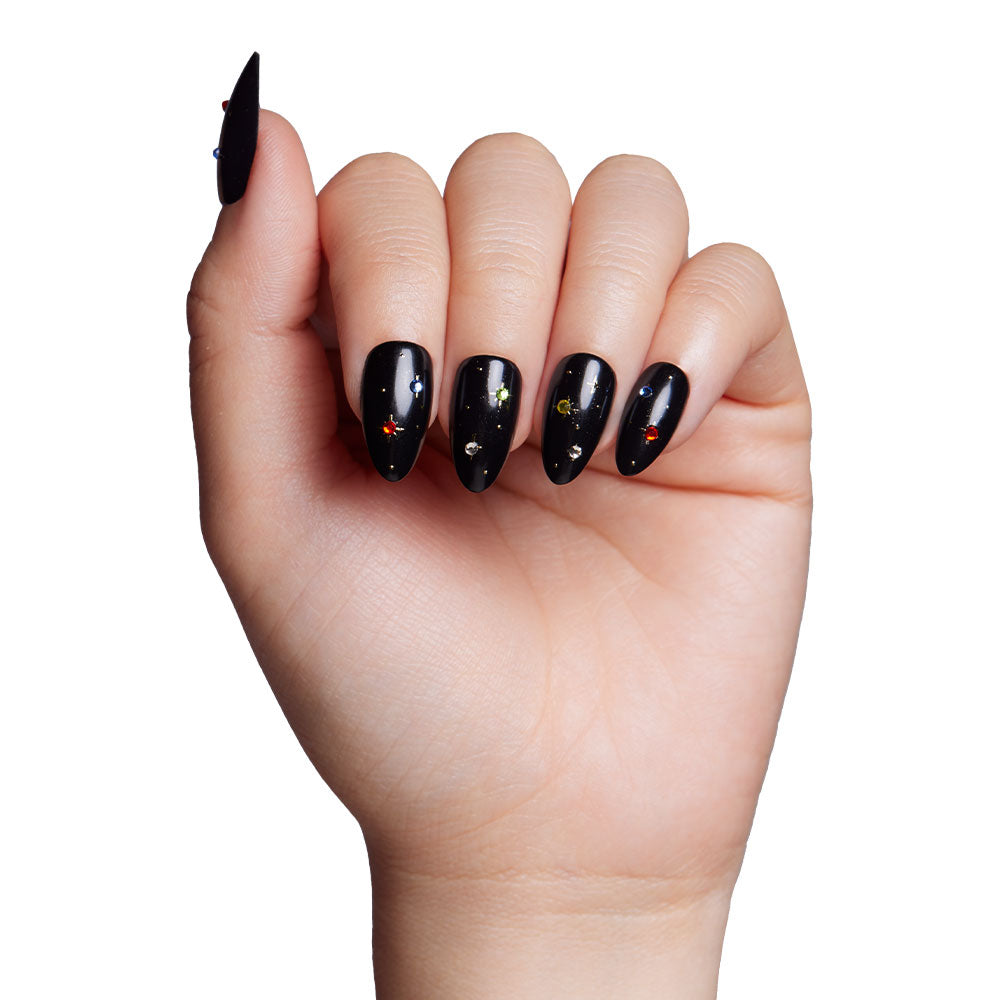 A hand displays glamnetics Winter Comet: glossy black, long almond nails adorned with colorful celestial stars and dots. Fingers are slightly curled against a white background. - Product image