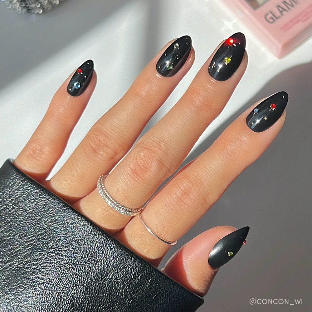 A hand with glamnetics Winter Comet almond nails—black with colorful dots—plus three slim silver rings, is shown against a softly lit background. - Product image