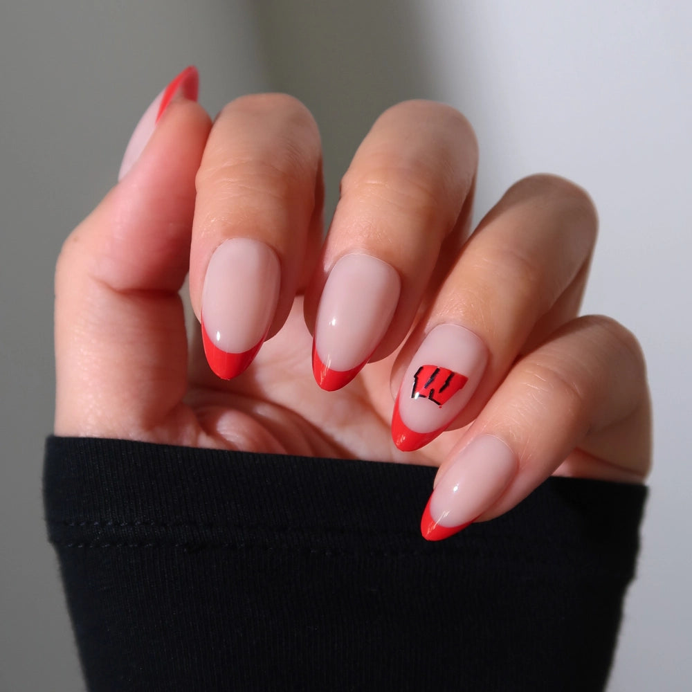 A hand with short almond nails shows a nude base and bold red tips, with the ring finger sporting a black and red W—styled after the glamnetic Wisconsin Badgers Press-On Nail Set. The person is wearing a black sleeve. - Product image