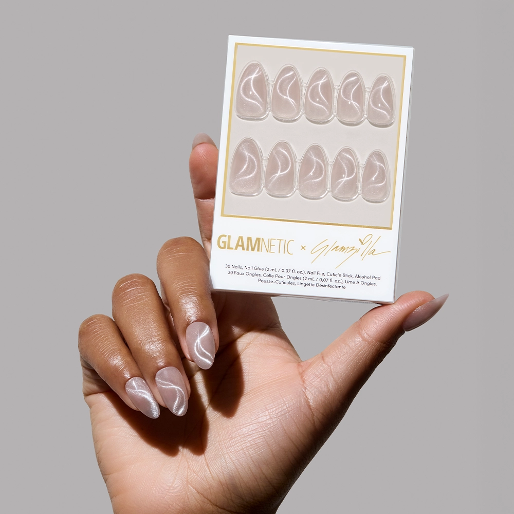 A hand holding a Glamnetic x Glamzilla press-on nail kit showing 10 almond-shaped nails with a nude shimmer base and white swirl accents, labeled with gold branding.