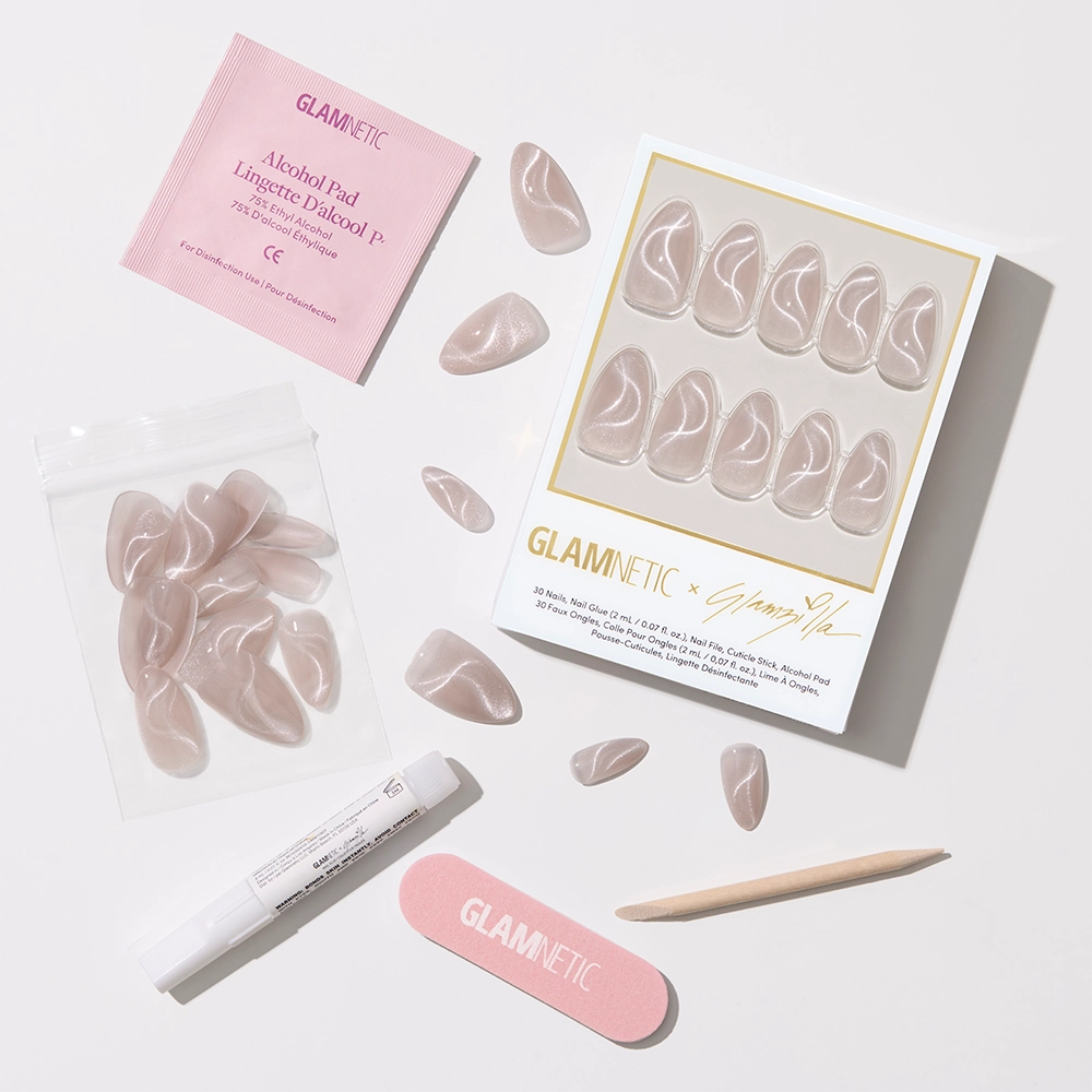 Flat lay of the Glamnetic x Glamzilla “Woo-Man!” press-on nail kit, featuring 30 nude shimmer nails with white cat-eye swirl designs. Surrounding items include a pink Glamnetic nail file, wooden cuticle stick, nail glue, alcohol prep pad, and a resealable bag of extra nails — all arranged neatly on a white background. - Product image