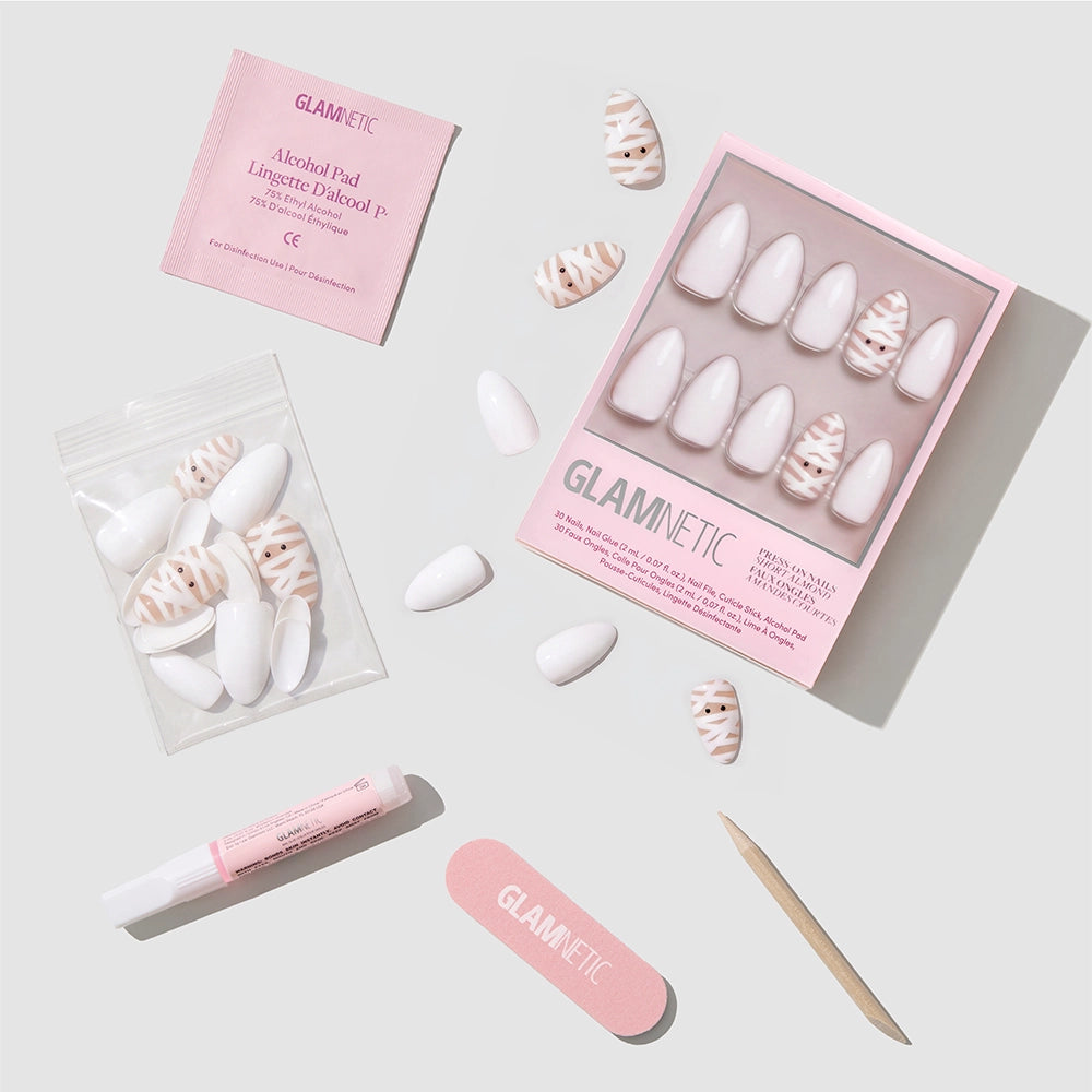 The glamnetic Wrapped Up set features short almond press-on nails in pink and white patterns, highlighted by a chic white mummy nail design. The kit includes nail glue, a nail file, a wooden stick, an alcohol pad, and extra nails—all presented on a light background. - Product image