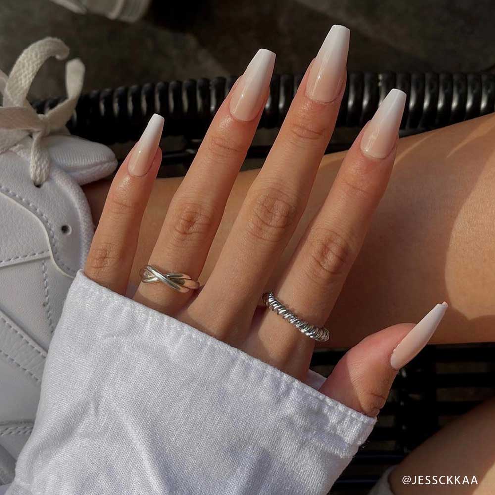 A hand with long, customizable coffin-shaped Creme de Nude ombre acrylic nails by glamnetic, featuring a glossy finish and two silver rings, rests on a lap. The person wears a white long-sleeve shirt and white sneakers are in the background. - Product image