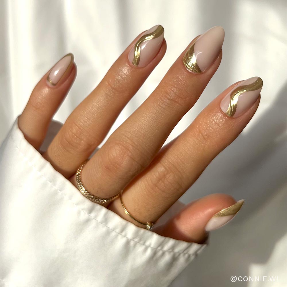 A hand with short, almond-shaped nails flaunts the glamnetic Material Girl set: a nude base with abstract gold foil swirls. The look is paired with two delicate gold rings and a white long-sleeved shirt for added chic style. - Product image