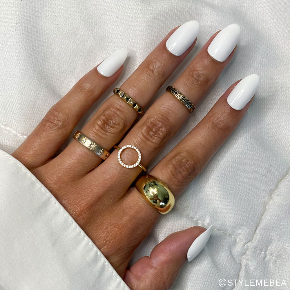 A hand wears glamnetic Angel long, white, almond-shaped press-on nails and five textured gold rings. The person is dressed in a white long-sleeved shirt against a soft white fabric background. - Product image
