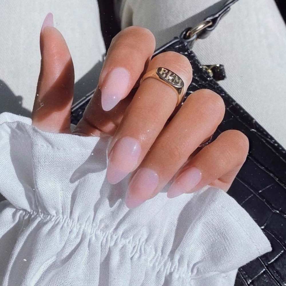 A hand with long, almond-shaped nails in light pink—Cloud 9 by glamnetic—rests on a textured black bag. A gold ring and ruffled white sleeve complete the look, highlighting a chic and elegant manicure. - Product image