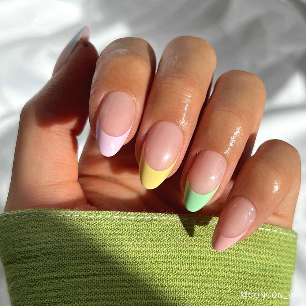 A hand with neatly manicured almond-shaped nails featuring the Confetti pastel tips from glamnetic in pink, purple, yellow, and green rests on a surface, complemented by a light green sleeve. - Product image