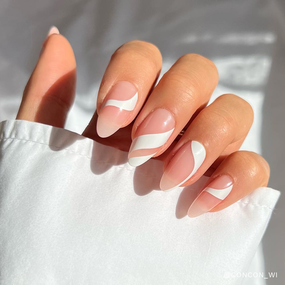 A hand with elongated medium almond-shaped nails, painted with glamnetic’s Marshmallow Swirl press-ons—nude base and white wavy lines. Nails are partially covered by a white sleeve and illuminated by bright natural light. - Product image
