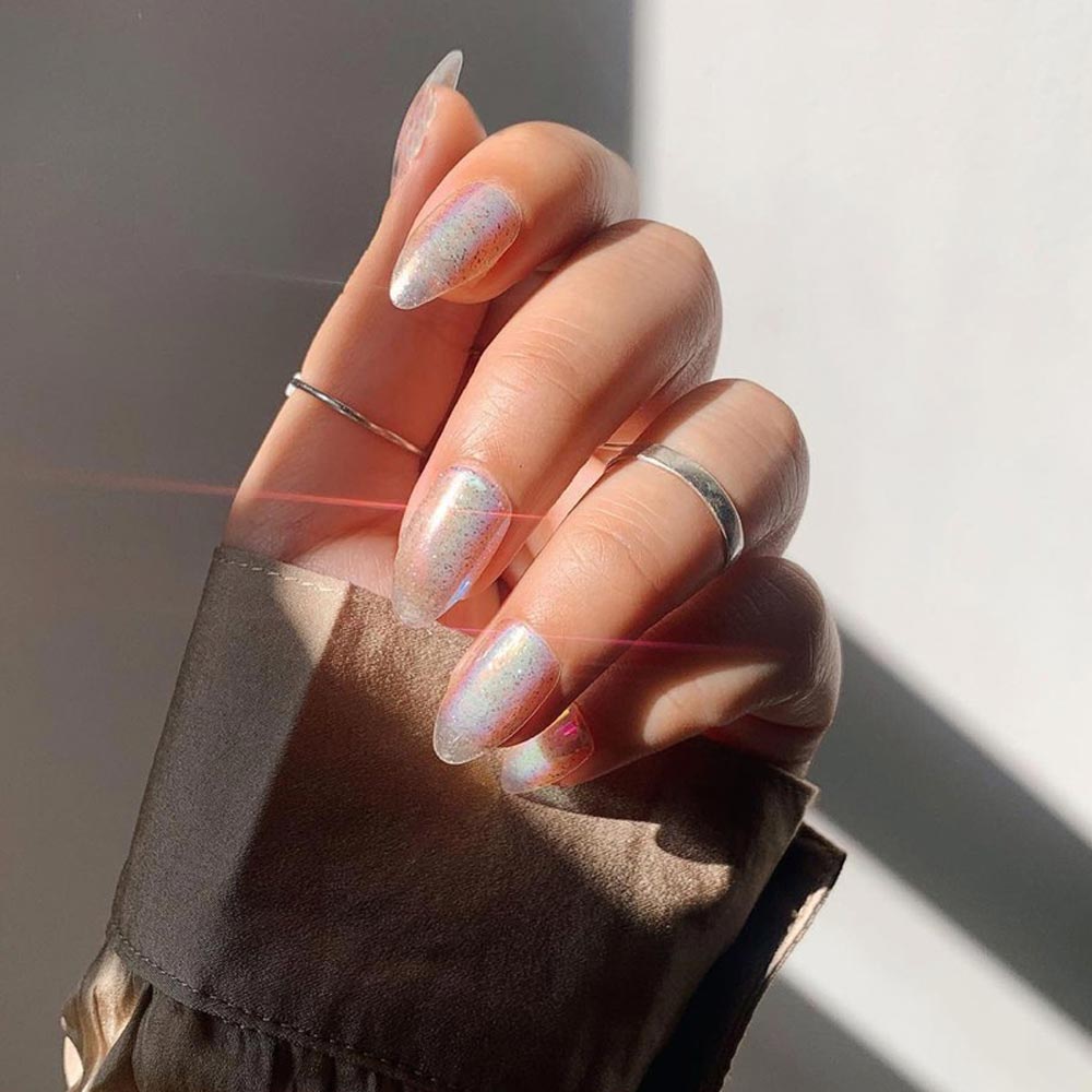 A hand with long, pointed almond nails painted in glamnetics Stardust—shiny, iridescent polish. Silver rings and a brown satin long-sleeve shirt complete the look as sunlight highlights the holographic finish. - Product image