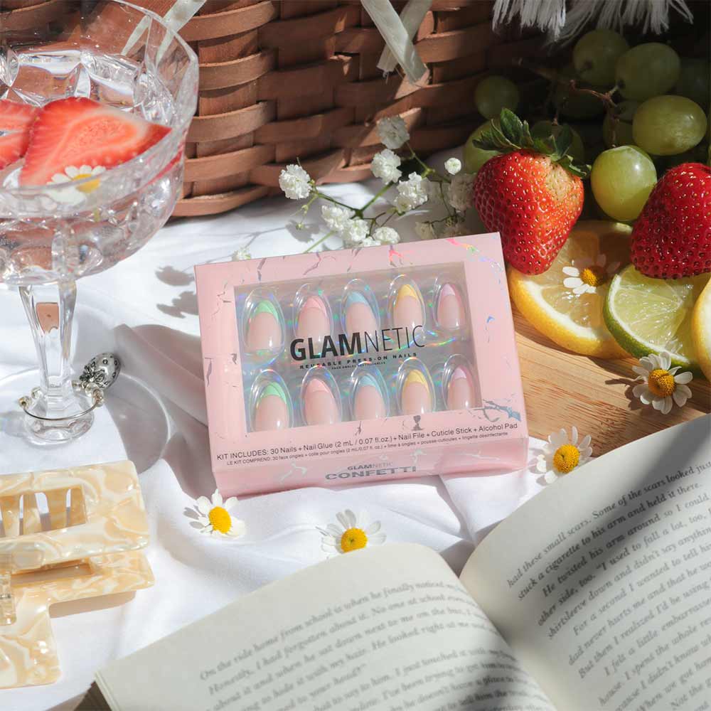 A box of glamnetic Confetti press-on nails with pastel tips and almond shape sits on a white cloth next to strawberries, lemon slices, strawberry water, hair clips, and an open book in the sunlight. - Product image