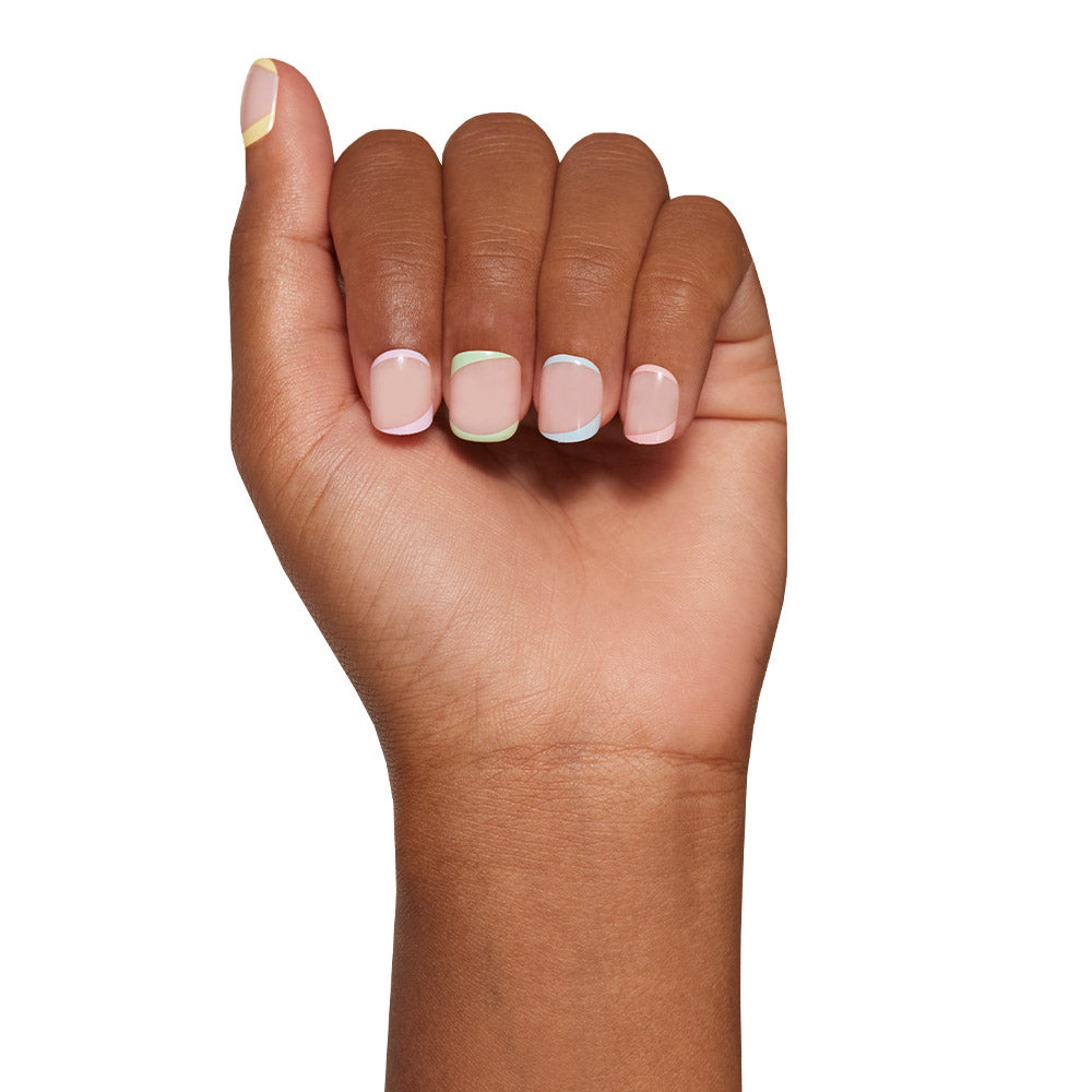 A hand with super short, neatly manicured nails painted in glamnetic’s Tutti Frutti, a pale nude set with thin, colorful pastel tips on each nail, shown against a white background. - Product image