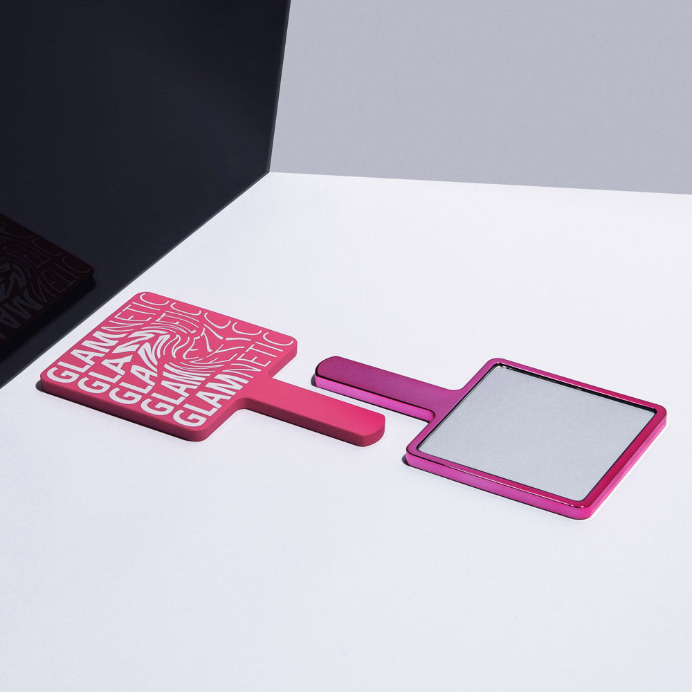 Two pink glamnetic Handheld Mirrors sit on a white and dark gray surface. One mirror, face down, shows the brand name; the other stands upright, ideal for use as a lash application mirror to check your look. - Product image