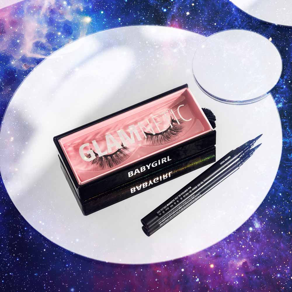 A box of glamnetic Babygirl vegan false eyelashes and two black liquid eyeliners are displayed on a mirrored surface with a galaxy background. These reusable lashes create bold, glamorous looks. - Product image
