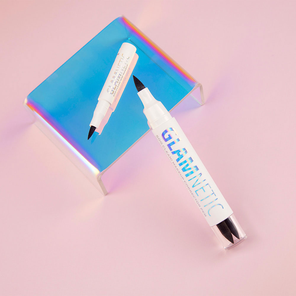 Two white glamnetic Soo Clean! Duo dual-tip eyeliner pens with Vitamin E stand upright, reflected on a shiny iridescent surface, against a soft pink background. - Product image