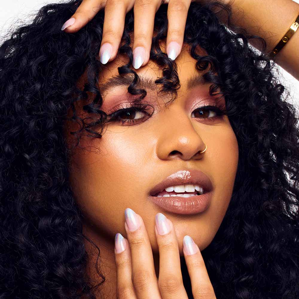 A woman with curly dark hair poses, showing off shimmery pink eyeshadow, glossy lips, and pastel almond-shaped nails with a semi-transparent finish using glamnetic’s Winter Storm. She wears a gold nose ring and bracelet. - Product image