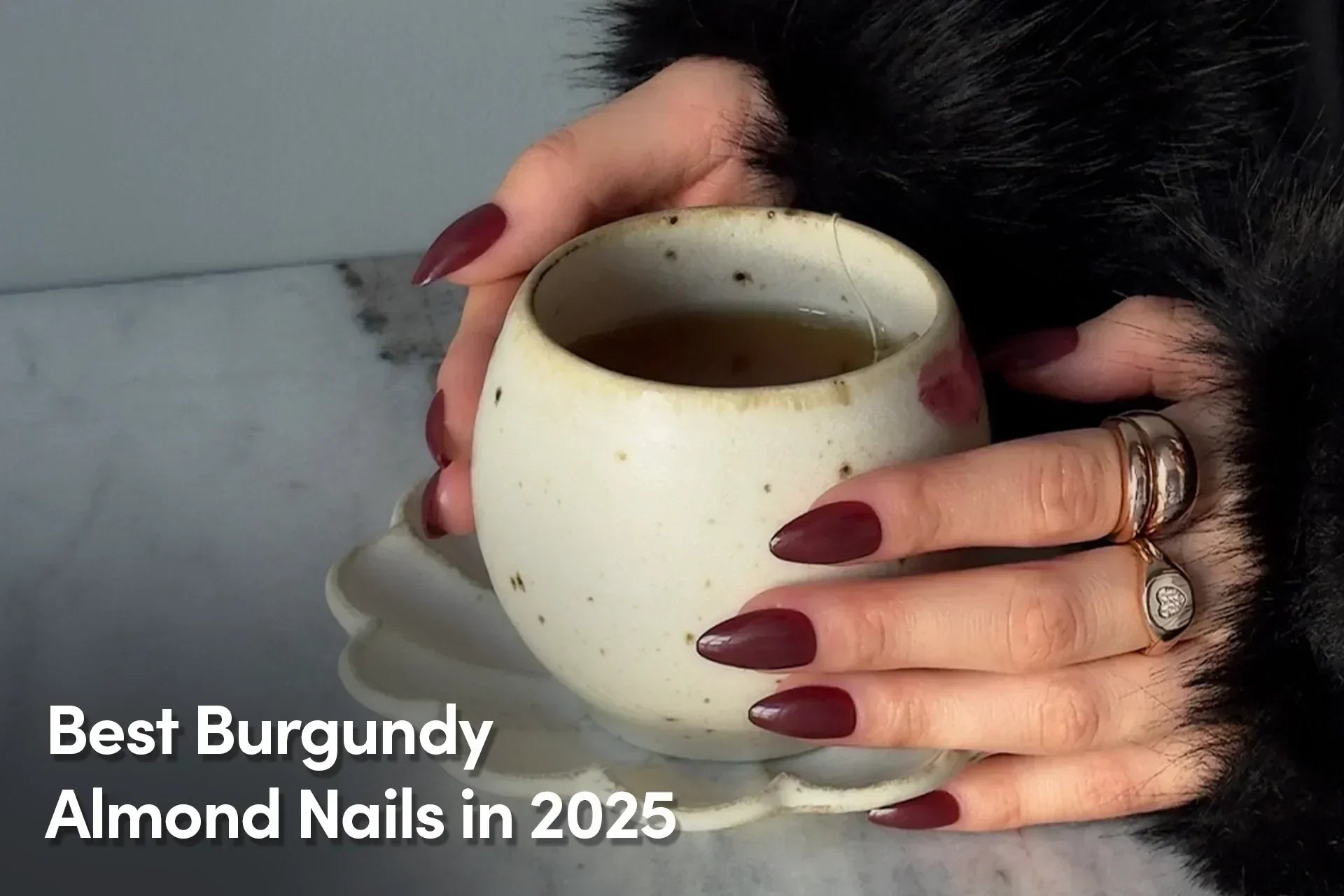 Two hands with burgundy almond-shaped nails holding a handmade ceramic cup, accessorized with chunky gold and silver rings and a faux fur cuff.