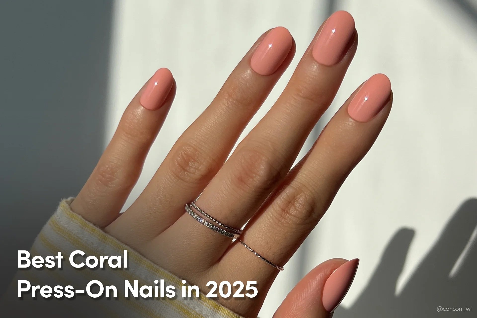 Hand with short, rounded coral press-on nails, wearing delicate silver rings, and a soft yellow striped sleeve visible