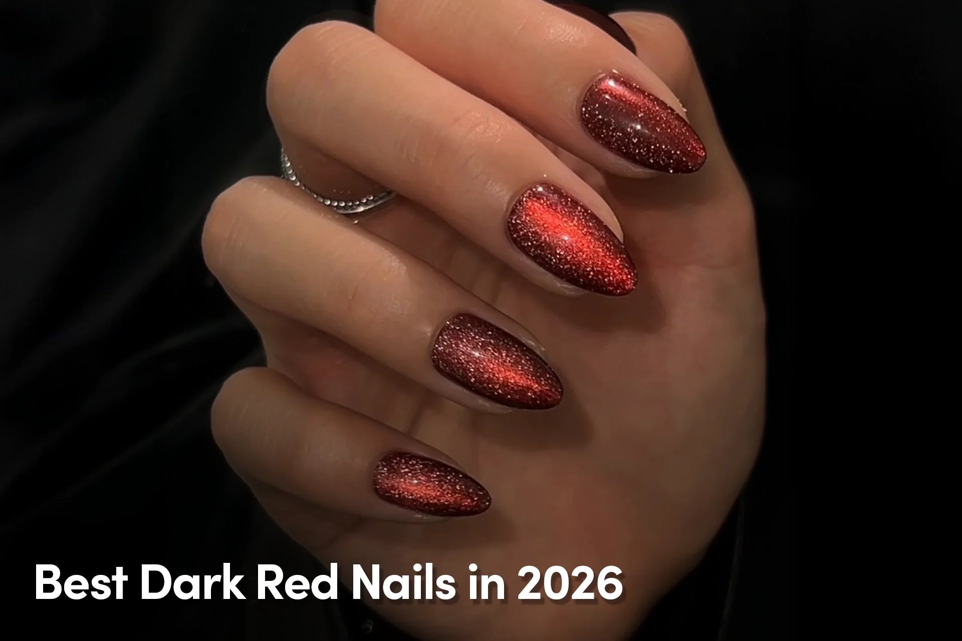 Hand with shimmering dark red metallic almond-shaped nails with a sparkling finish against a black background.