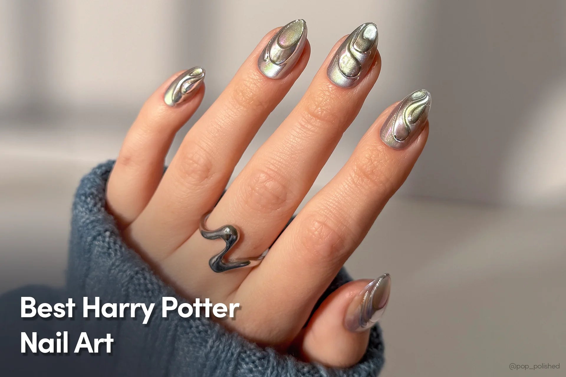 A hand with reflective silver chrome Harry Potter–inspired nails, shown against a soft background while wearing a snake-shaped ring and a blue sweater.