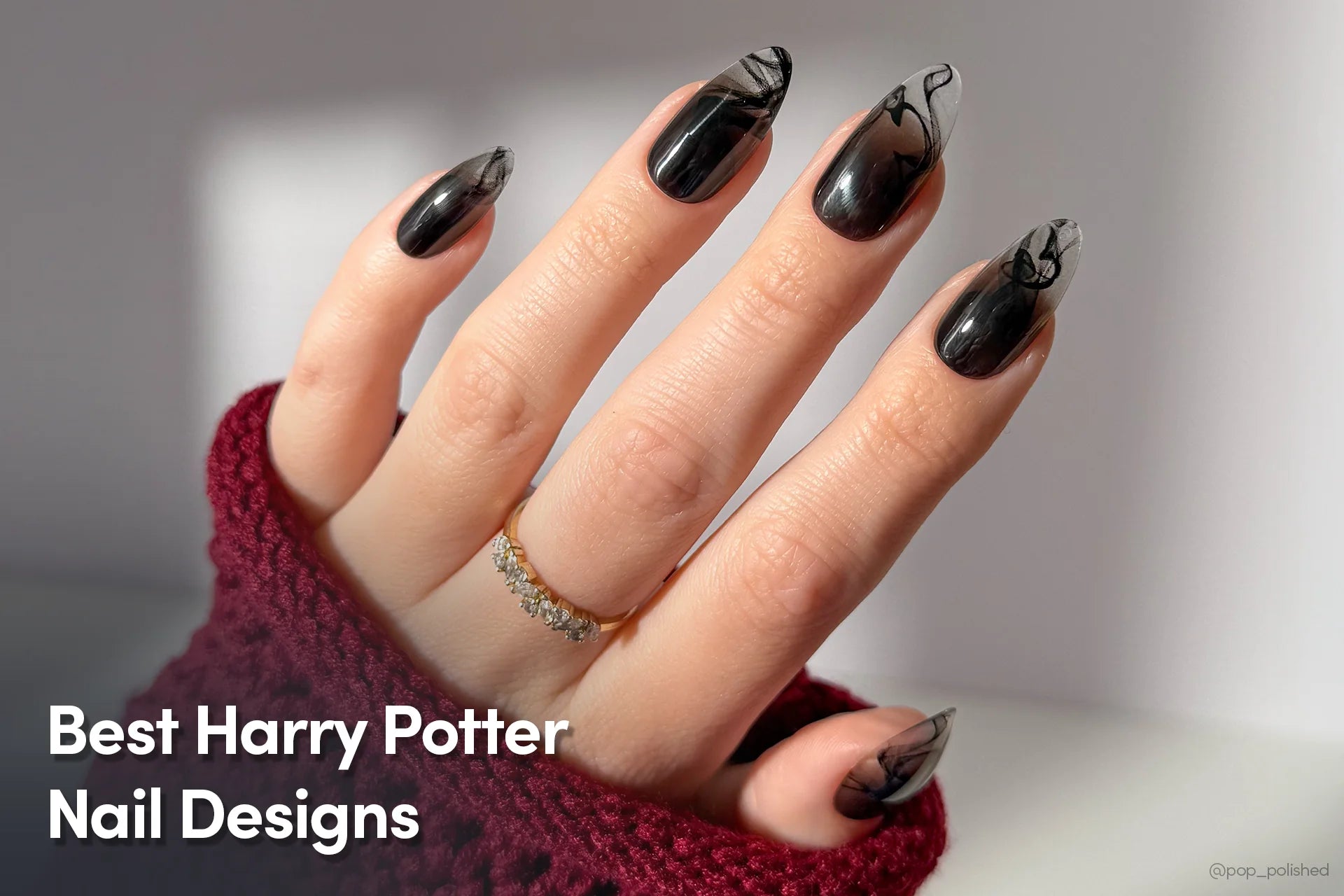 A hand with long, black marble-patterned Harry Potter–inspired nails, shown against a soft background while wearing a burgundy sweater and a gold ring.