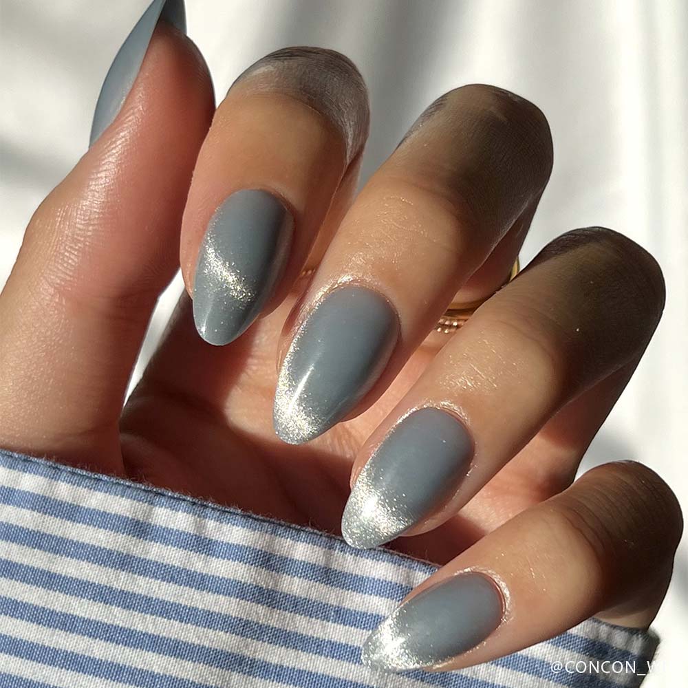 A hand with almond-shaped nails painted in glamnetics Baby Blues—matte gray with shiny silver tips and a velvet metallic finish—rests against a blue and white striped shirt, soft sunlight highlighting the look. - Product image