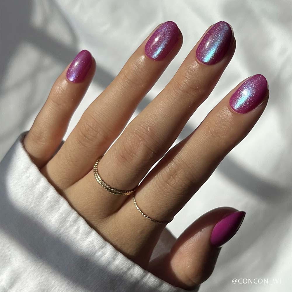 A hand with glamnetics Berry Fizz iridescent magenta nails and a velvet metallic finish, adorned with gold rings, rests against a softly lit background. The person is wearing a long-sleeved white top.