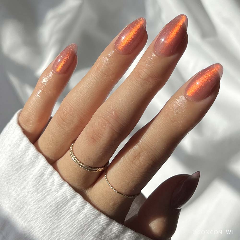 A hand with long, almond-shaped nails painted in glamnetics Rosé—a shimmery, iridescent orange velvet metallic polish—wears two gold rings and a white shirt against a softly lit background.