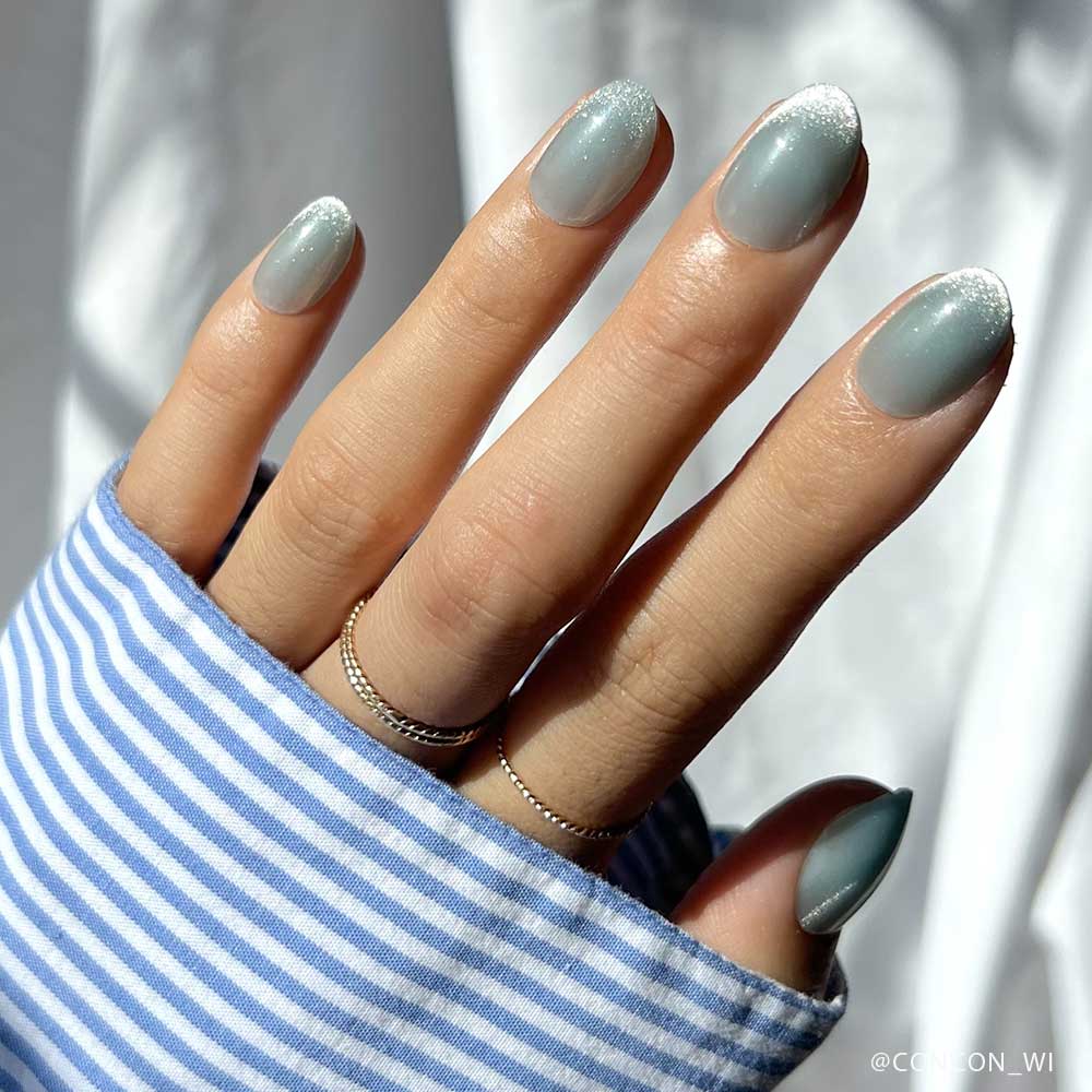A hand with short oval nails in glamnetic’s Unicorn Tears, a silver-blue ombré, is shown in natural light with blue and white striped sleeves and silver rings, highlighting the velvet metallic nails effect.