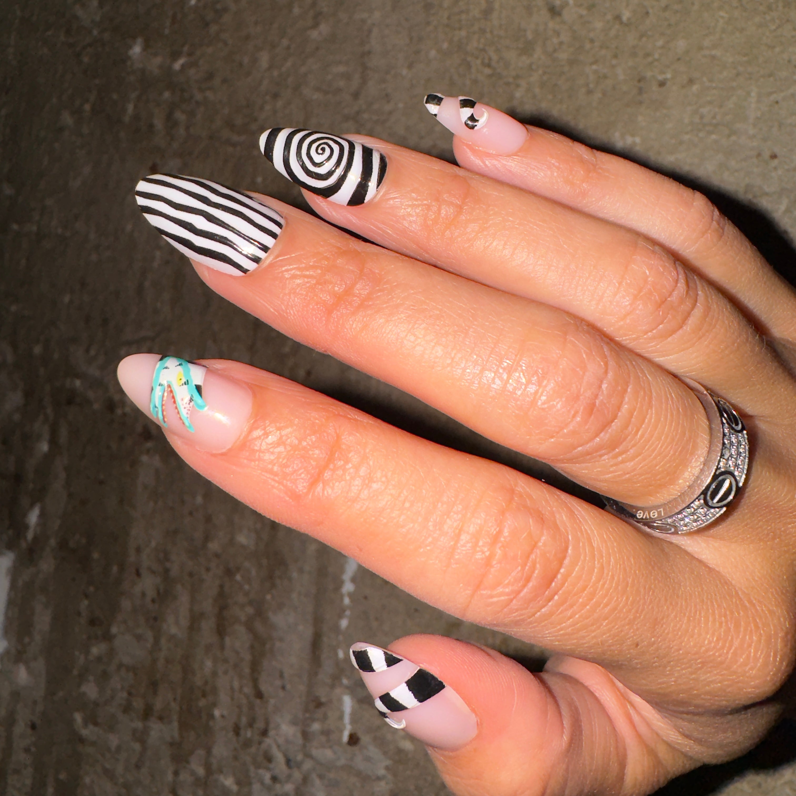 A hand with almond-shaped nails wears the glamnetic Sandworm set, each nail showing unique black and white stripes, spirals, checks, and minimal art. Two silver rings complete the look against a textured background. - Product image
