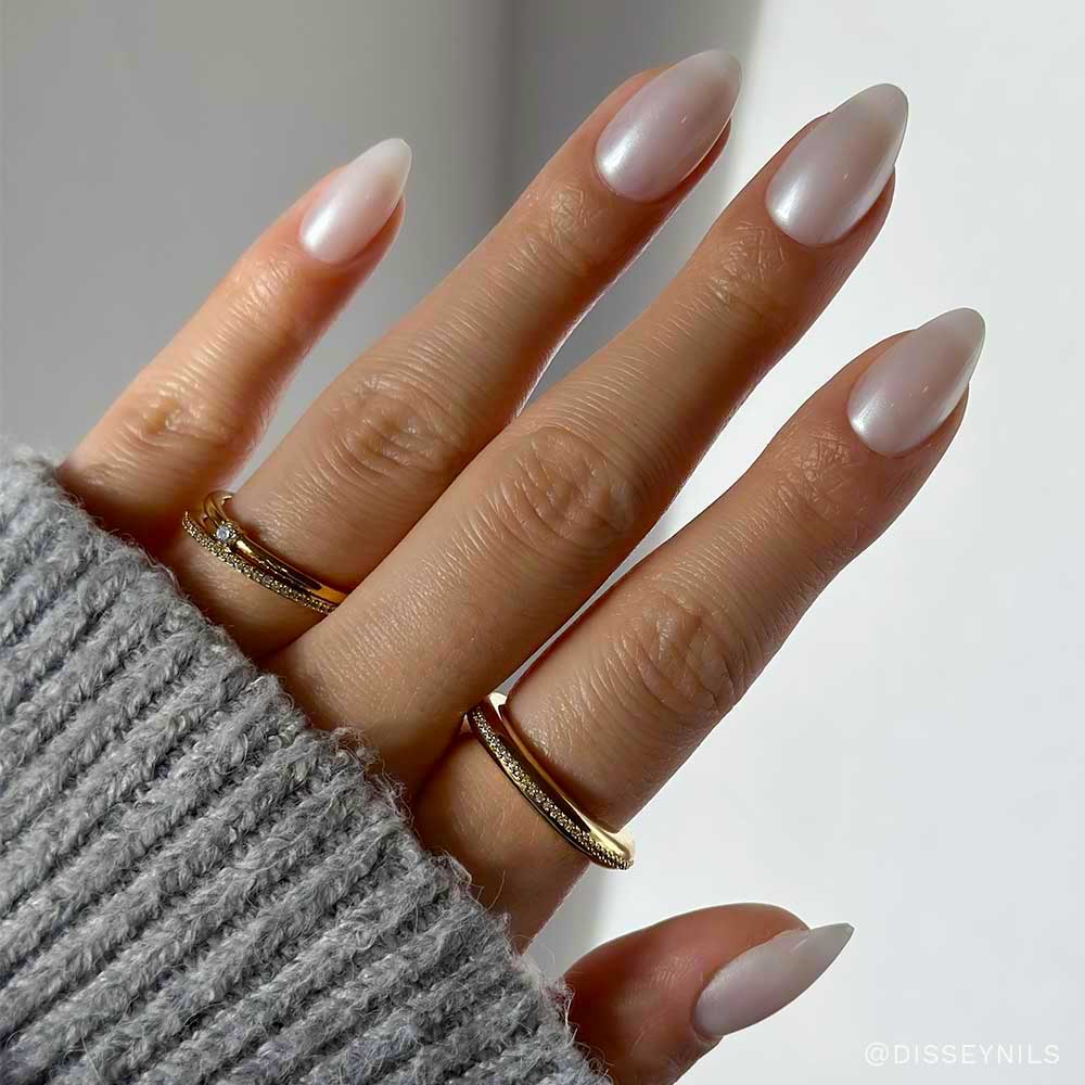 A hand with almond-shaped, glossy, pale pink glamnetic press-on nails from the Nail Lover's Gift Set, wearing two gold rings on the middle and ring fingers and a light grey knitted sweater. - Product image