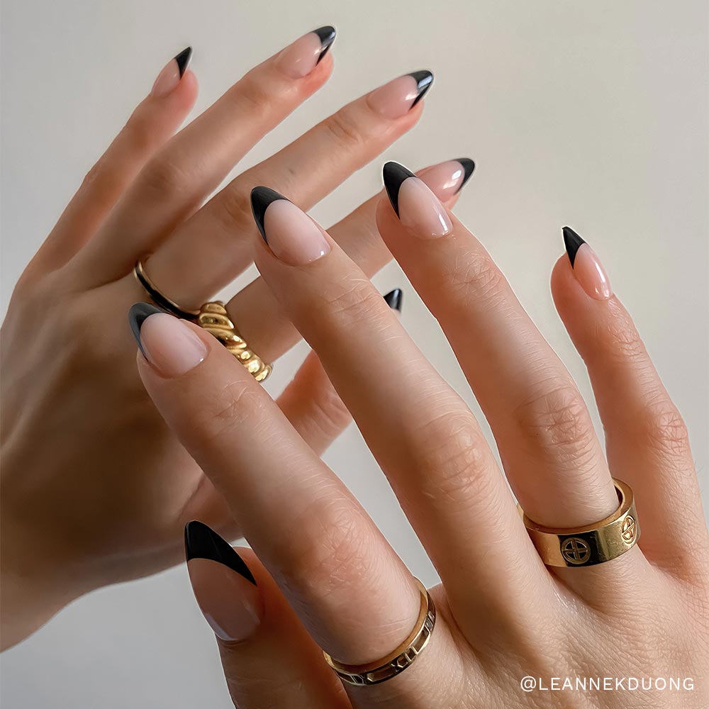 Close-up of hands with almond-shaped press-on nails from glamnetic's Nail Lover's Gift Set, featuring a nude base and black tips. Gold rings accent the look, while a neutral background highlights this chic nail set. - Product image