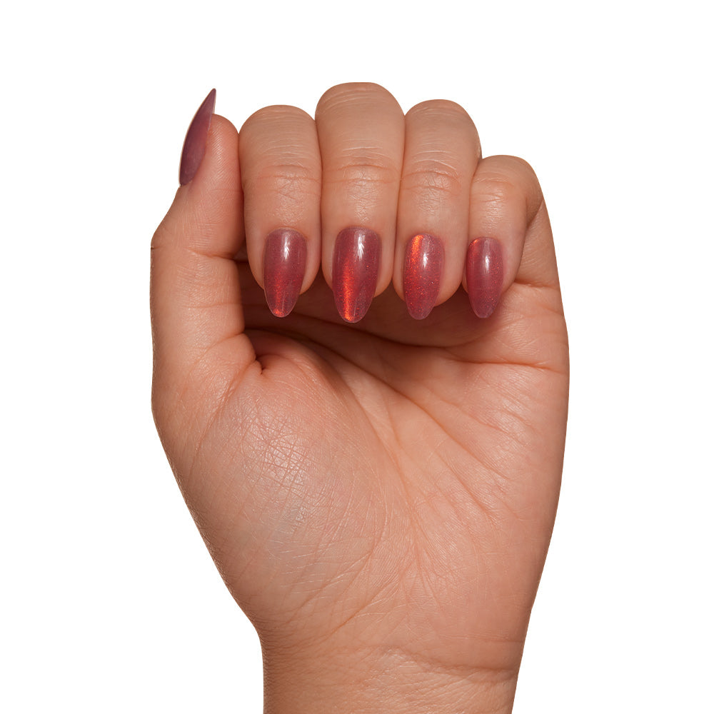 A hand with medium skin tone showcases glamnetic’s Rosé Nail Set, featuring long almond-shaped nails in a shimmery reddish-pink velvet metallic finish, displayed against a white background. - Product image