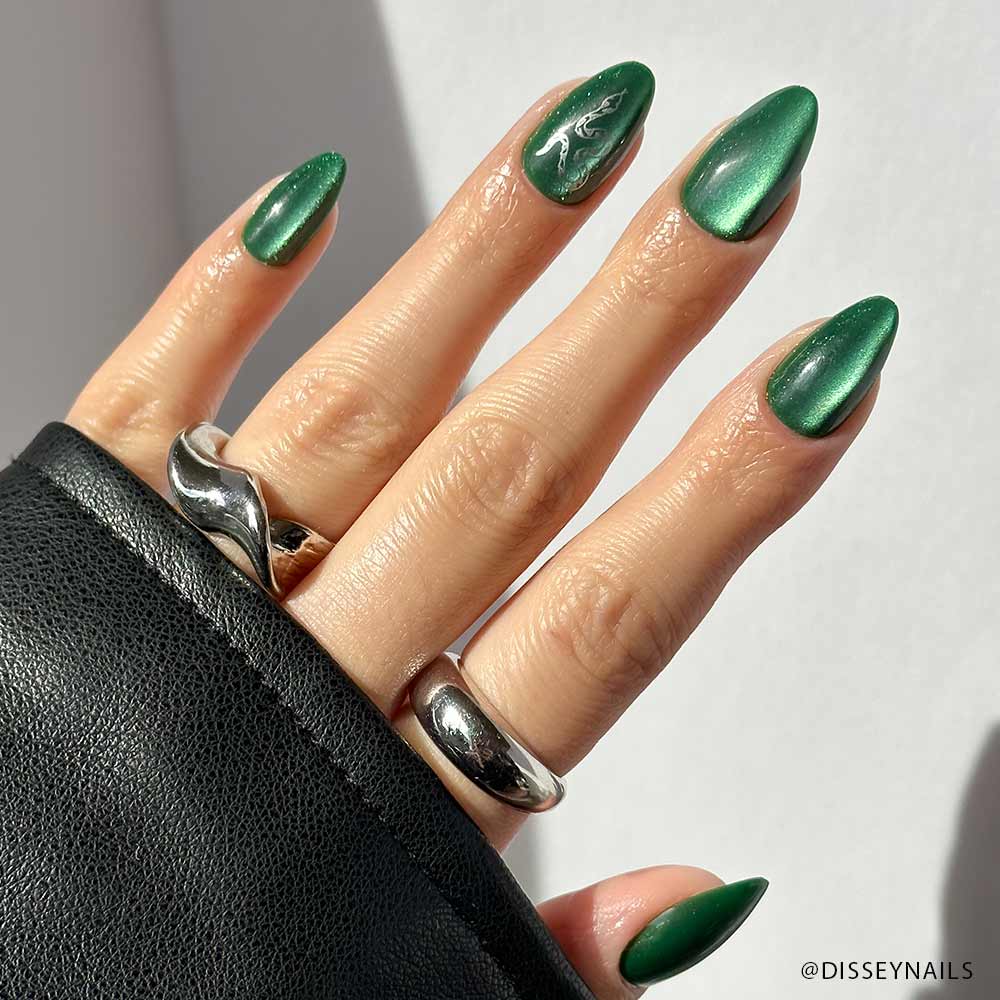A hand sports almond-shaped, emerald green Slytherin™ nails by glamnetic—one with a subtle alligator design. Chunky silver rings decorate the fingers, partially covered by a black leather sleeve.