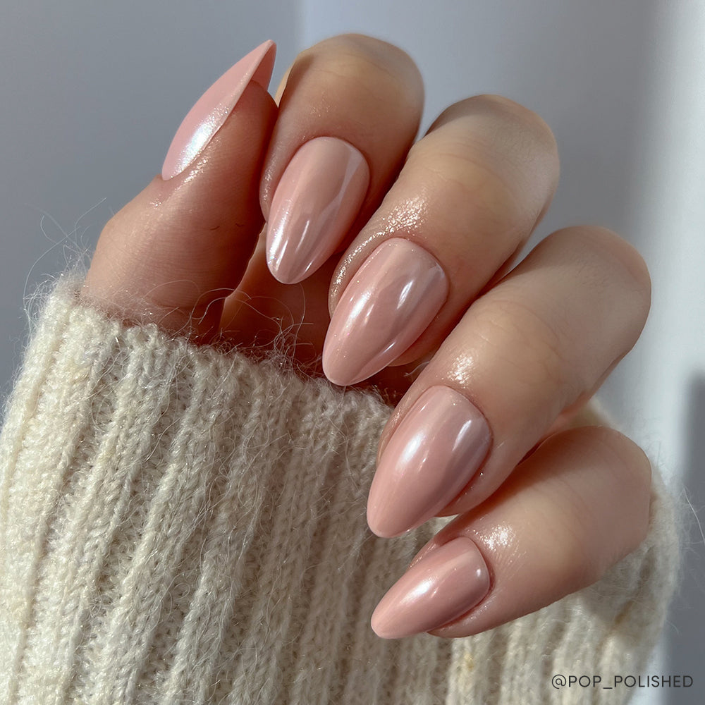 A hand with short almond-shaped nails in a glossy blush pink, possibly using the glamnetic First Kiss set, is shown against a soft light background. The person is wearing a cream-colored knit sweater. - Product image