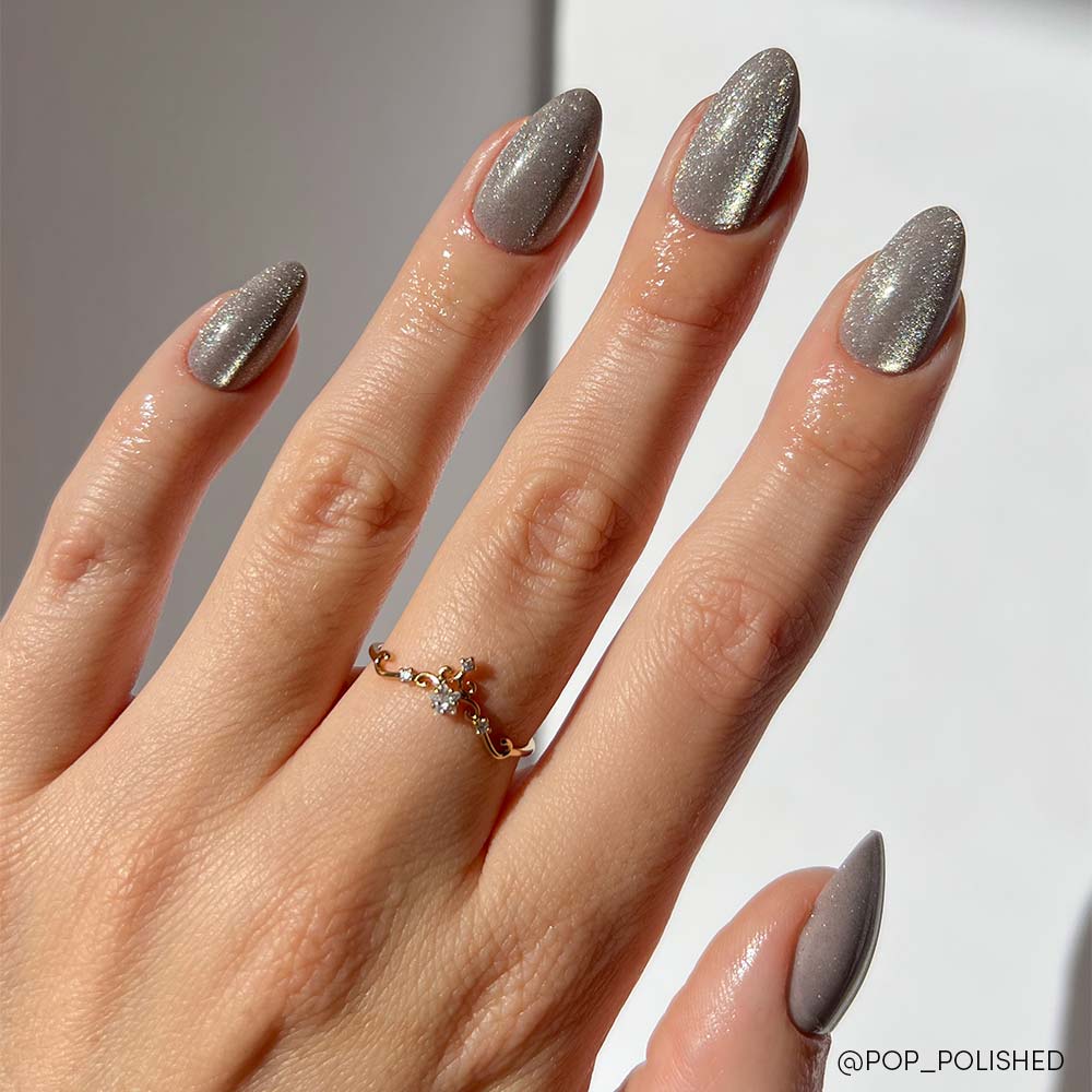A hand with short-almond nails painted in glamnetic’s Oslo, a shimmery silver cat-eye, wears a delicate gold ring with small gemstones. The fingers are slightly spread against a plain, light background. - Product image