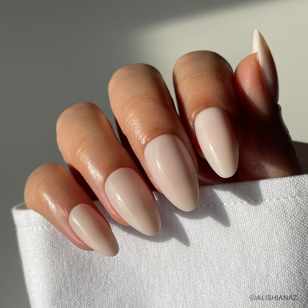 A hand with short almond-shaped nails painted in glamnetics glossy Ballet Pink set rests on white fabric, shown in natural sunlight.