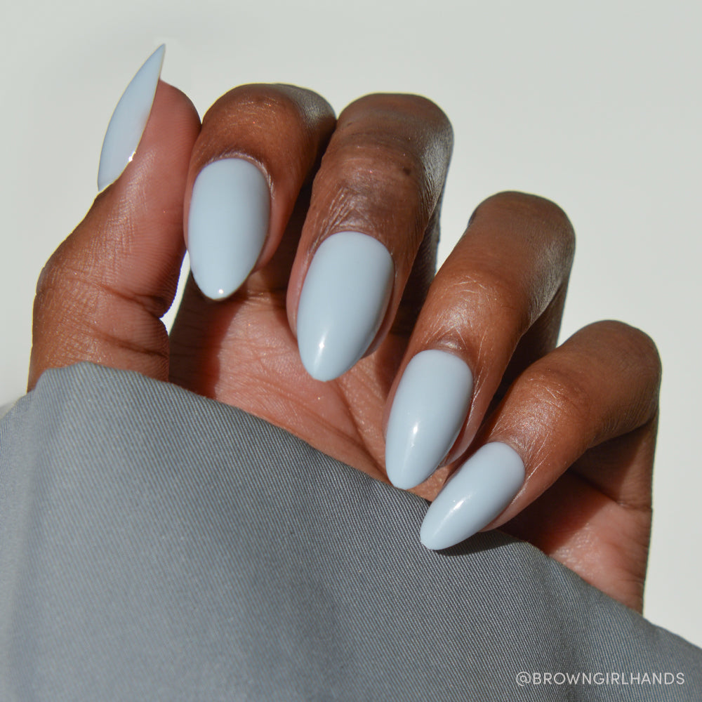 A hand with dark skin tone shows off glamnetic’s Blueberry Milk press-on nails in a glossy finish on short almond-shaped nails, partly covered by a gray sleeve.
