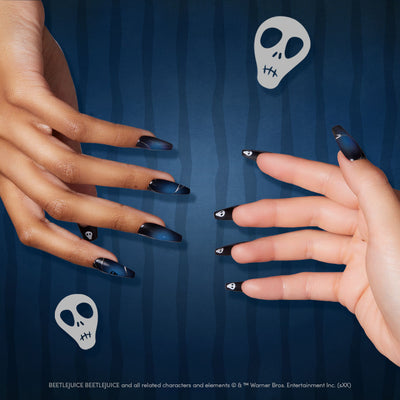 Two hands wearing Glamnetic x Beetlejuice press-on nails in a sleek dark blue and black gradient design, displayed over a striped blue background with white skull graphics