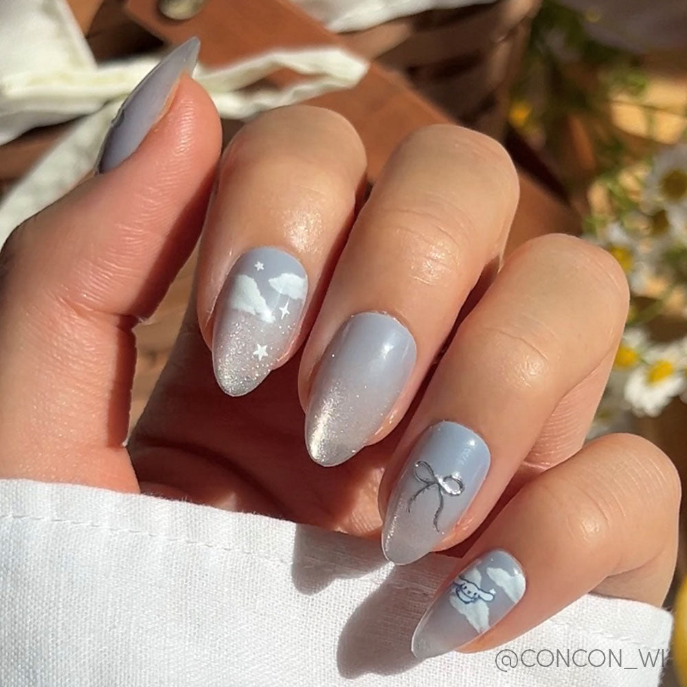 A hand with light blue and white gradient nails boasts delicate designs like a moon, stars, a bow, and a smiling face. Sunlight highlights the shiny glamnetic Cinnamoroll™ Cloud set with 3D silver accents for extra detail.
