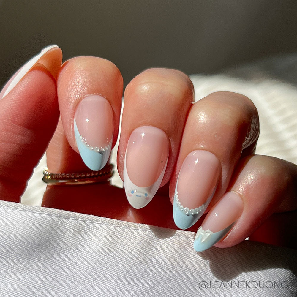 A hand with a Short Almond Design wears the glamnetic Cinnamoroll™ French Tip—pastel blue tips, tiny white beads, fine lines, and heart accents on a sheer nude base—resting on white fabric in natural light.
