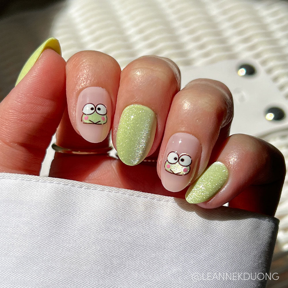 A hand with manicured nails shows off glamnetics Keroppi™ set, featuring light green glitter, solid yellow-green nails, and cute cartoon frog art. The background has a white textured surface and white clothing.