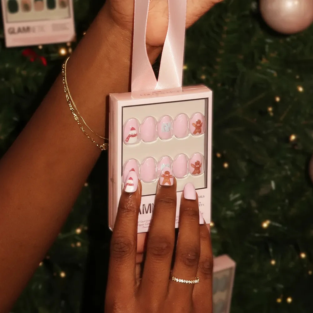 A person holds up a box of glamnetic Cocoa & Kisses, featuring pink festive short oval nails with candy canes and gingerbread men, in front of a Christmas tree. They accessorize with gold bracelets and a delicate ring. - Product image