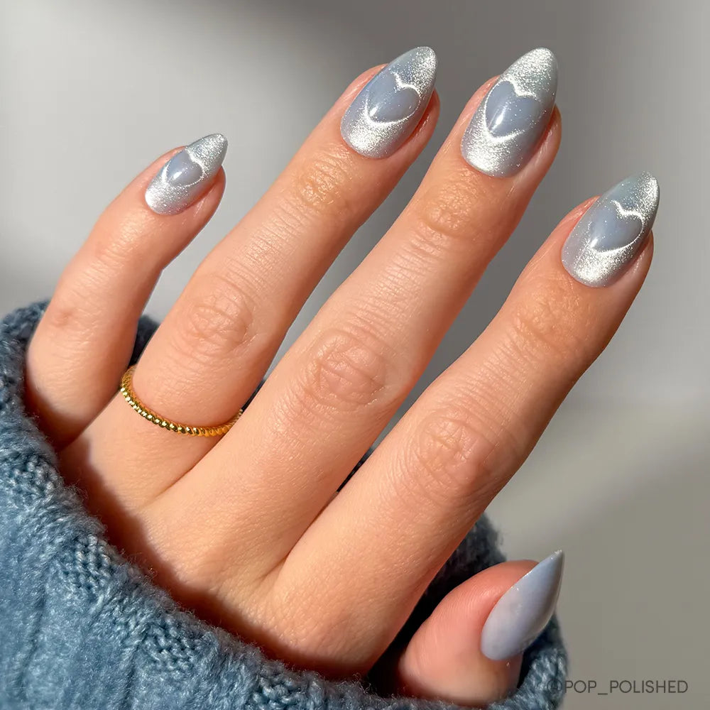 Short almond nails showcase shimmering silver polish with heart-shaped blue accents, inspired by glamnetic&#39;s Cold Hearted set. The wearer pairs them with a gold beaded ring and a cozy blue sweater.