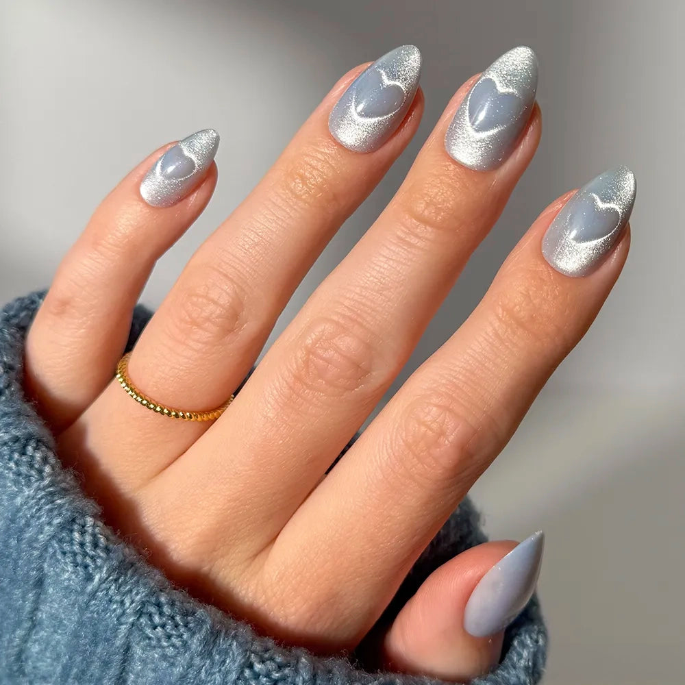 A hand with long, almond-shaped Cold Hearted nails by glamnetic—silver velvet cat-eye with blue heart designs—plus a gold beaded ring and a blue sweater sleeve partially covering the hand.