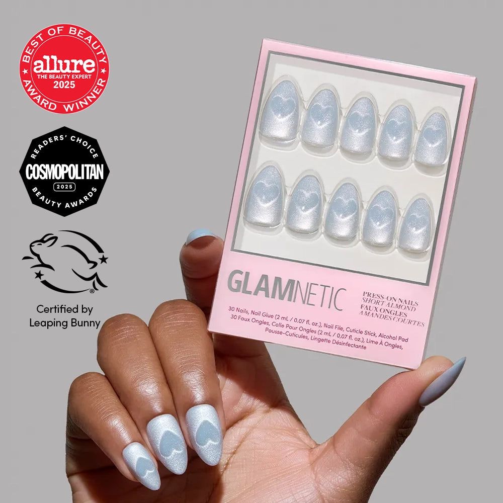 A hand shows off glamnetic Cold Hearted blue press-on nails with white clouds in a short almond shape, holding the package. Allure, Cosmopolitan, and Leaping Bunny logos appear on a neutral background.