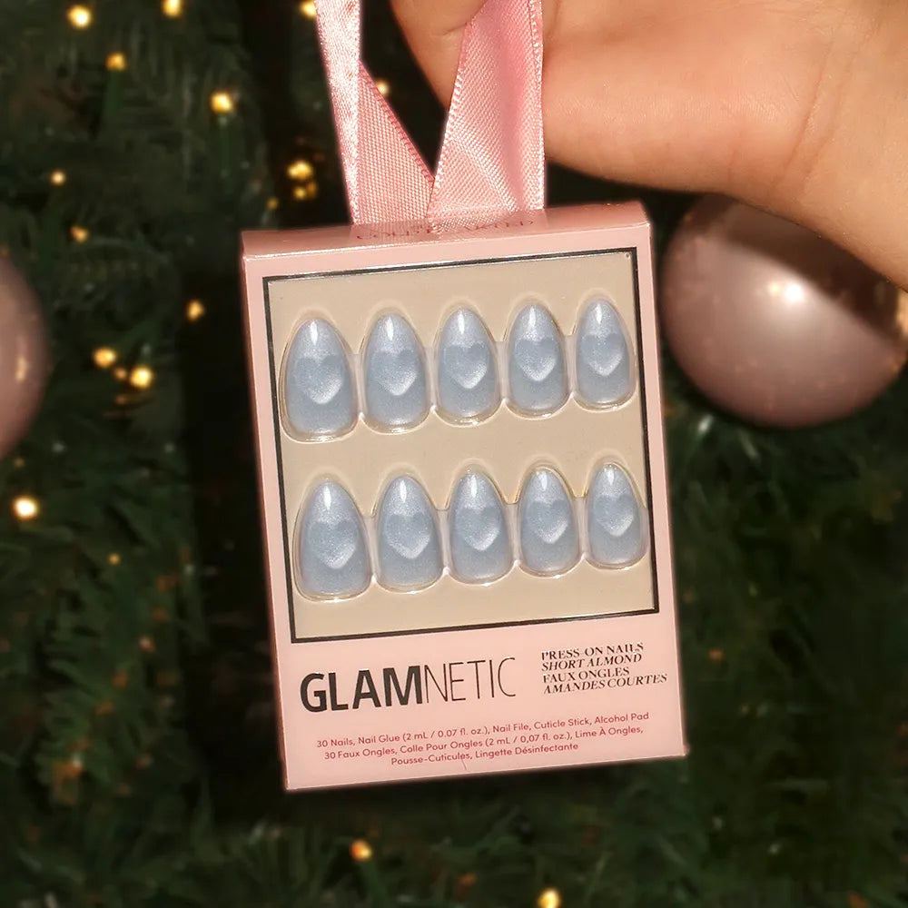 A hand holds a glamnetic Cold Hearted nail set with a pink ribbon in front of a Christmas tree decorated with pink ornaments; the short almond-shaped nails inside the box are metallic silver and neatly arranged. - Product image